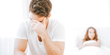 man covers face at foot of bed woman in background