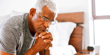 man sitting on edge of bed with hands folded thoughtfully