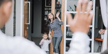 mother and young son holding hands at their front door and greeting his father