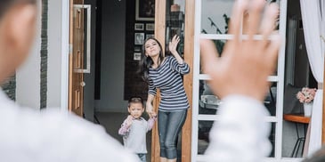 mother and young son holding hands at their front door and greeting his father