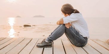 sad woman sitting on a pier looking out at a body of water at sunset