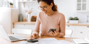 smiling woman using calculator and doing paperwork in front of her laptop