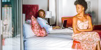 angry woman sitting at end of bed while husband works on his laptop in bed