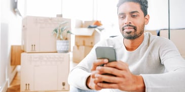 man looking at his phone in his home while surrounded by moving boxes