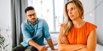 A man and woman sitting on bed separated with woman crossing arms upset