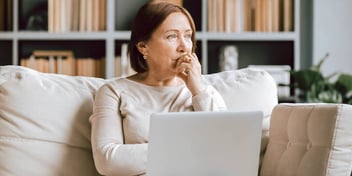 Older caucasian woman sitting on a coach working on her laptop staring off into the distance.