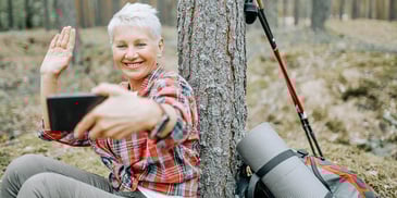 woman taking a selfie outside in the woods leaning against a tree by her yoga mat