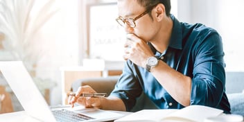 Young asian male working on his laptop conducting research with notebook open with pen in hand.