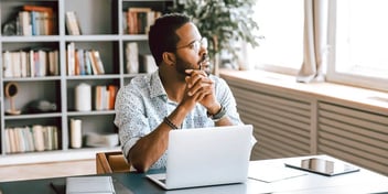 Young black male looking out of office window with laptop in front of him