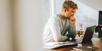 Young caucasian male sitting at desk working on laptop with notebooks around his desk.