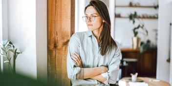 Young woman leaning against door frame staring out window with arms crossed